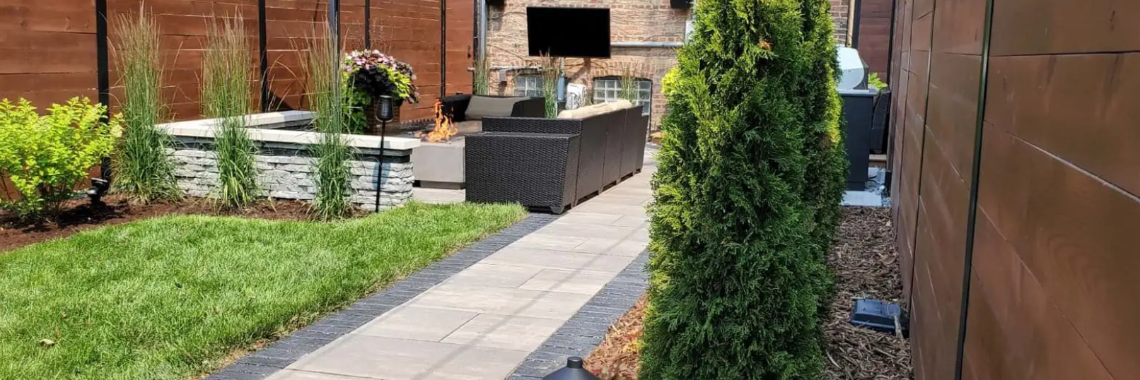 Backyard with a paved walkway, lawn, and garden beds. A tall green tree stands on the right. Brown fence in background.