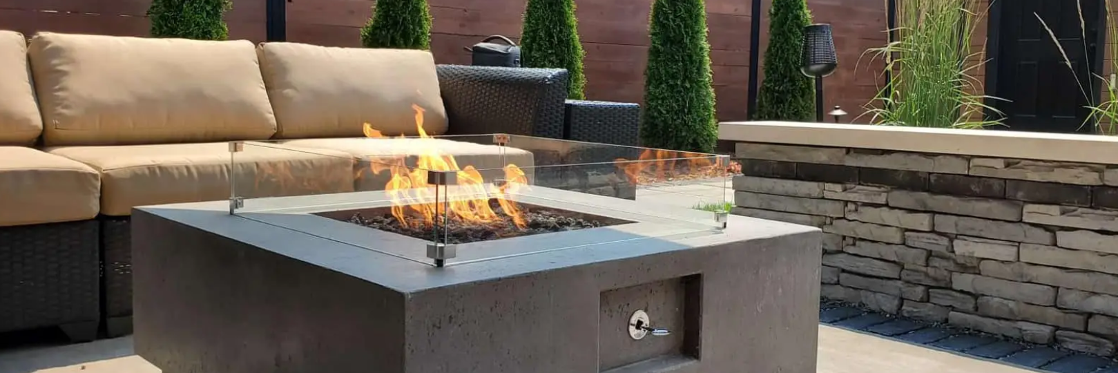 Outdoor patio with a lit fire pit surrounded by beige sofas. Tall green bushes and a stone wall complete the scene.
