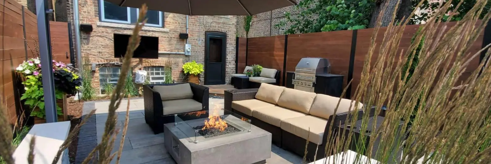 Outdoor patio with a fire pit, seating, and plants, enclosed by wooden fences and a brick building.