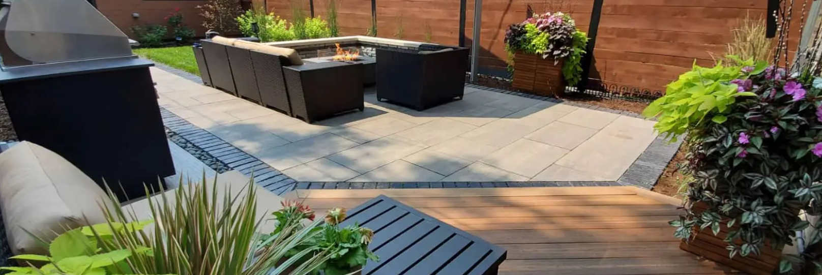 Backyard patio with dark furniture, plants, and a grill near a wooden deck.