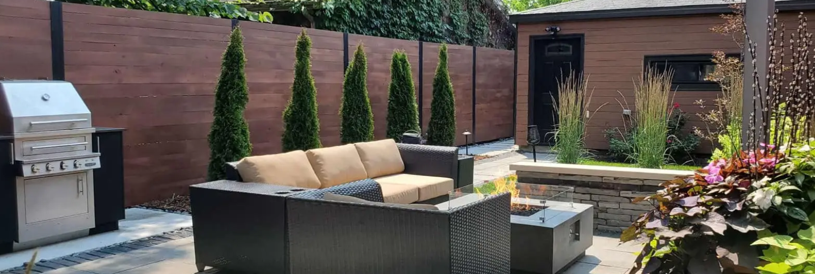 Outdoor patio with grill, sofa, fire pit, and decorative trees. Brown wood fence and a brick building in the background.