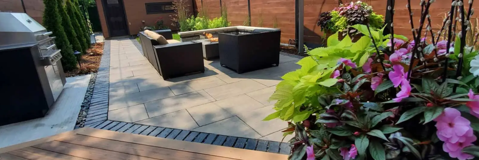 Patio with dark outdoor furniture, brick wall, lush plants, and a grill.