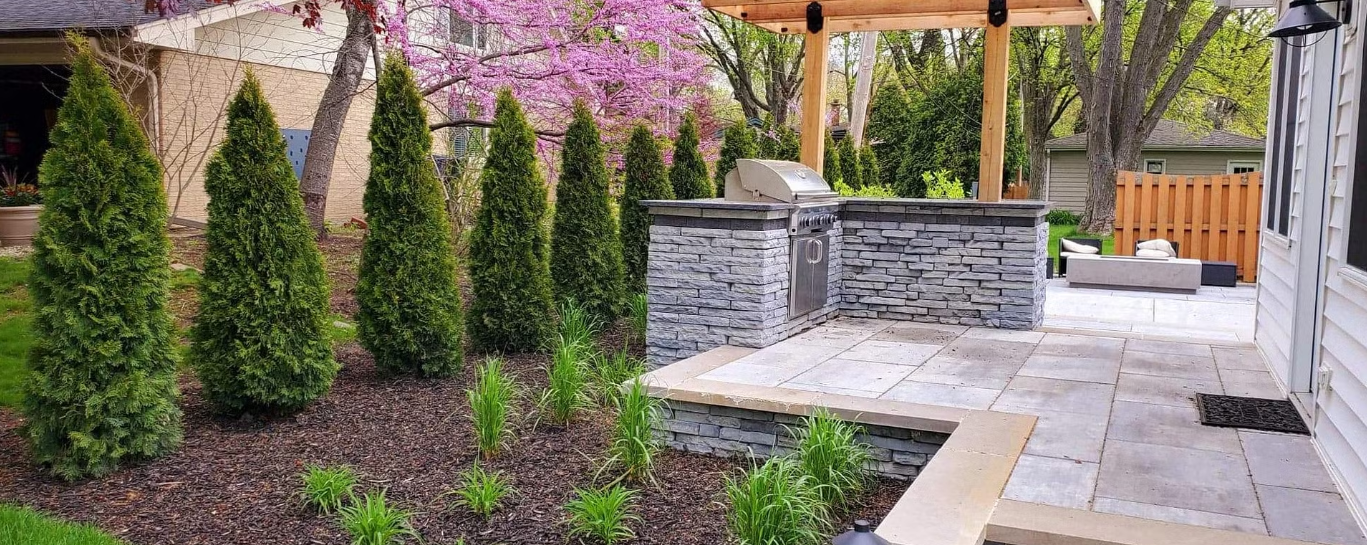 Backyard patio with grill, stone planters, green trees, and purple flowers.