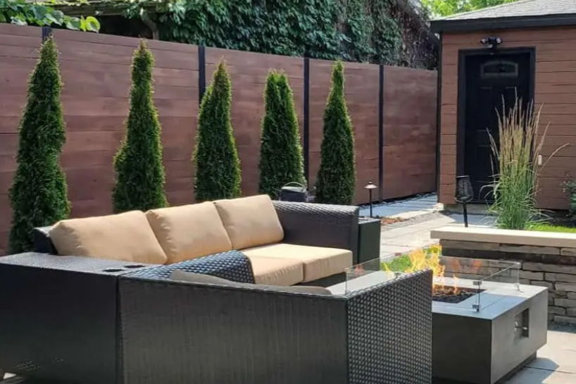 Outdoor patio with a brown sofa, a fire pit, and a wooden fence with trees.