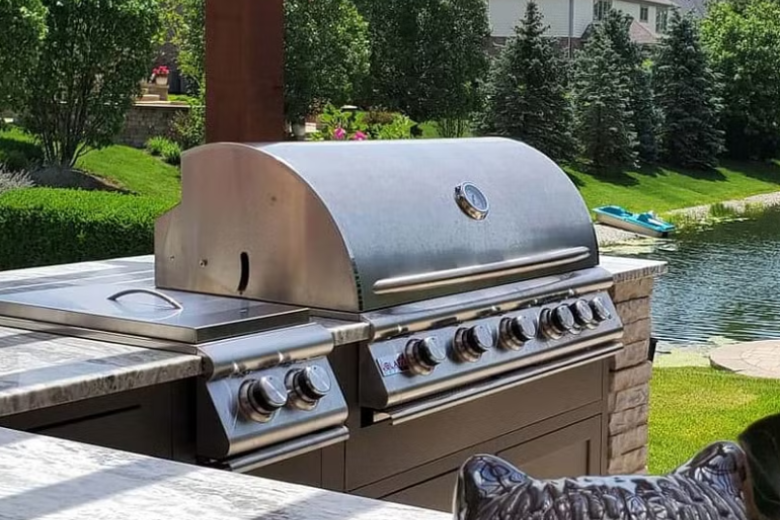 Stainless steel outdoor grill with closed lid on a countertop overlooking a lake.