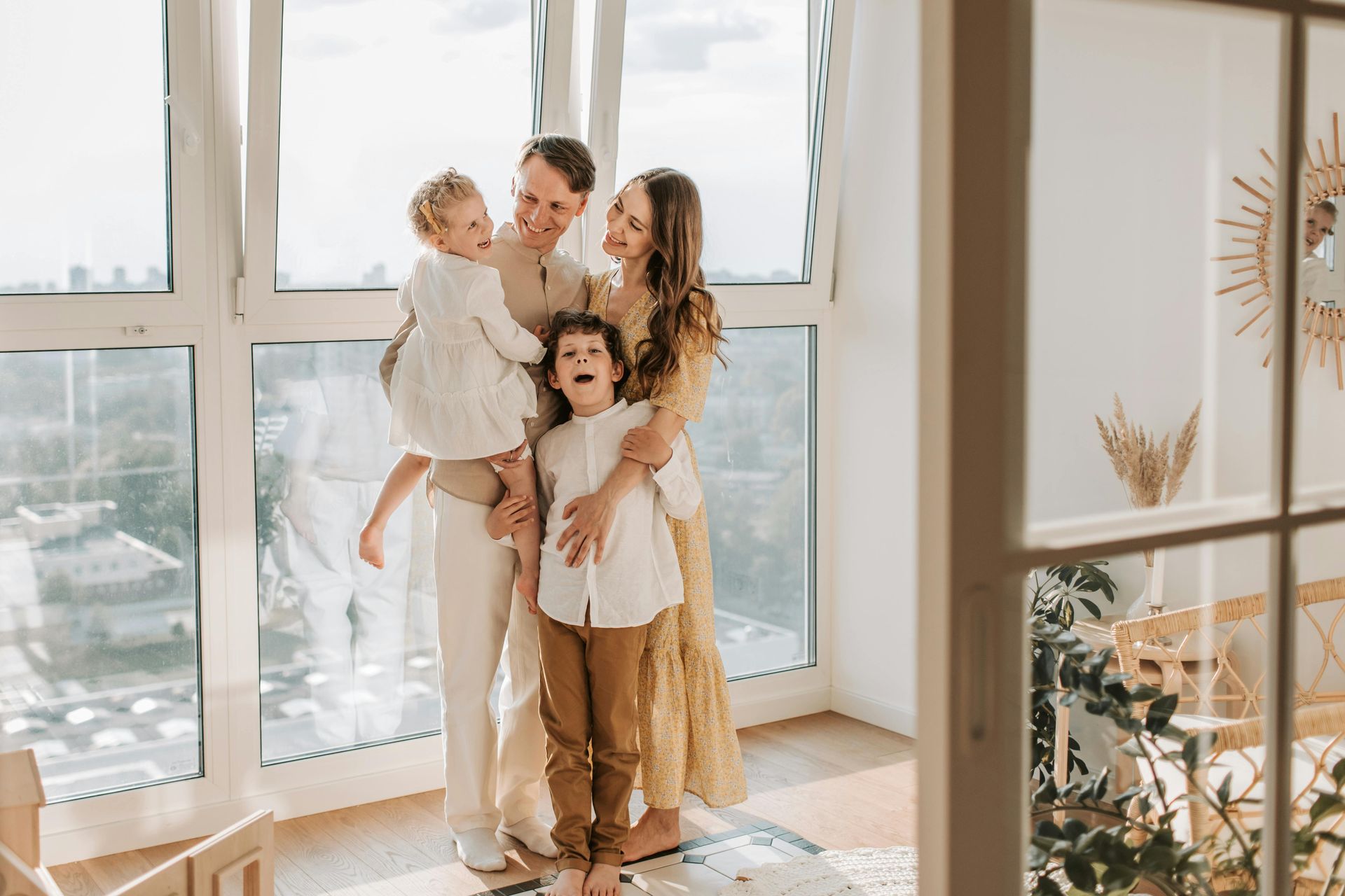 Family of four smiling by a large window; sunlit room. Woman embraces two children; man holds the youngest.