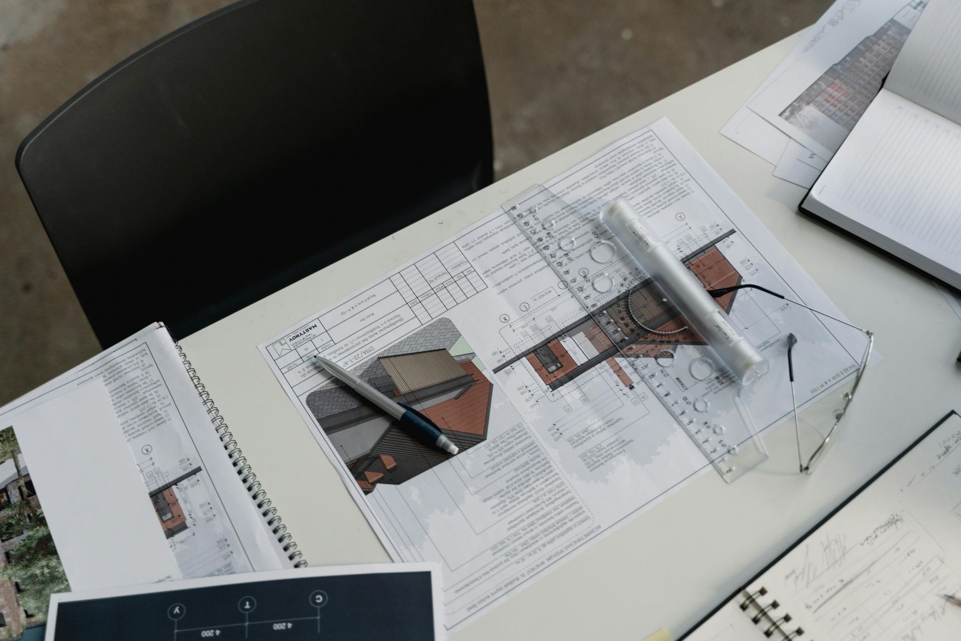 Architectural blueprints and sketches on a white table, alongside a pen, ruler, and notebook.
