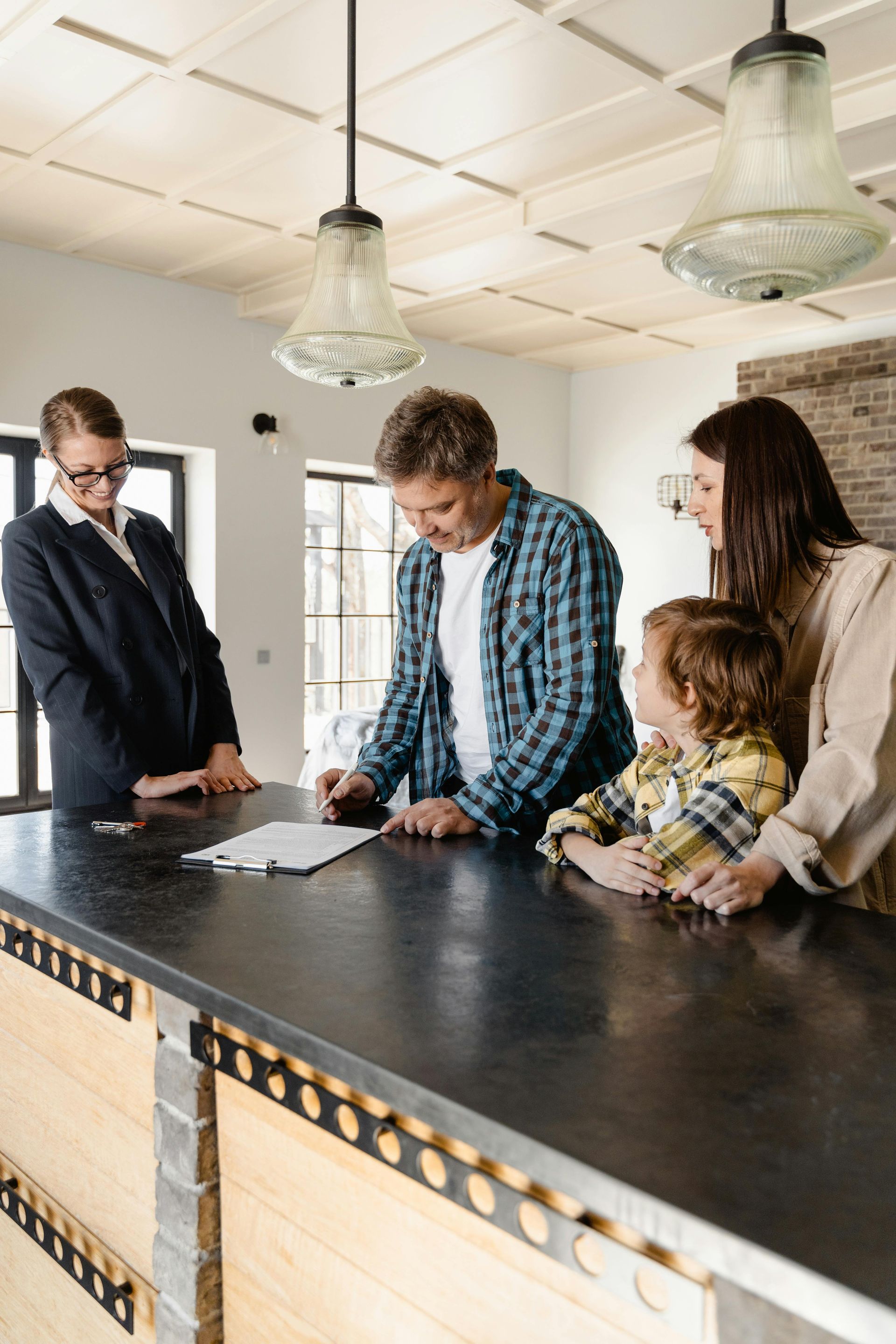 A family signs paperwork at a counter with a professional. The setting appears to be a home.