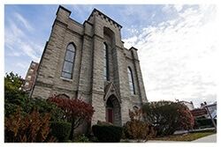 Gothic-style stone church with arched windows, a red door, and autumn foliage under a blue sky.