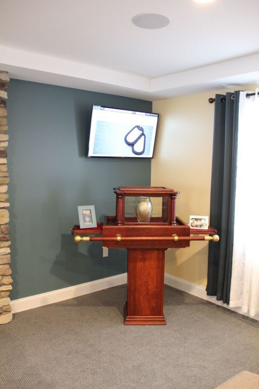 A wooden memorial table with a glass display holding an urn, under a mounted TV, in a corner of a room.