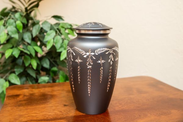 Black cremation urn with silver leaf design, sitting on a wooden table, greenery in the background.