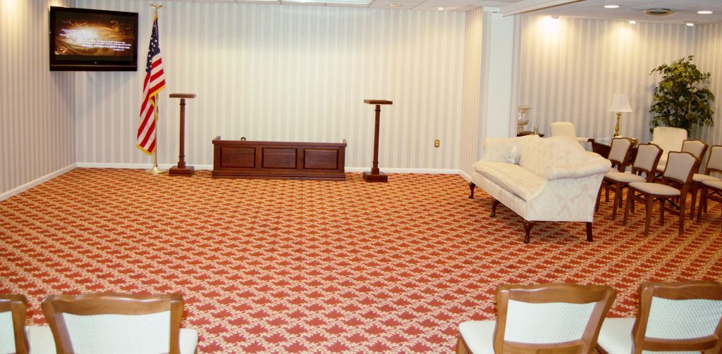 An empty, formal room with red patterned carpet and wooden chairs. An American flag stands.