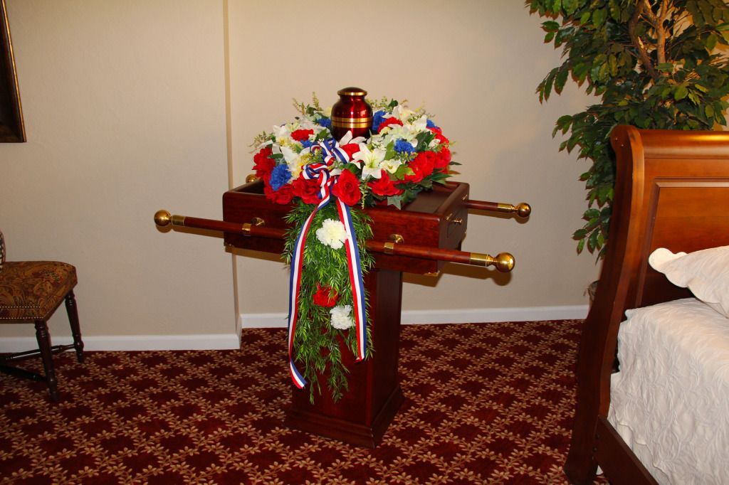 Cremation urn on display stand, adorned with wreath and patriotic ribbon, in a room with a bed and chair.