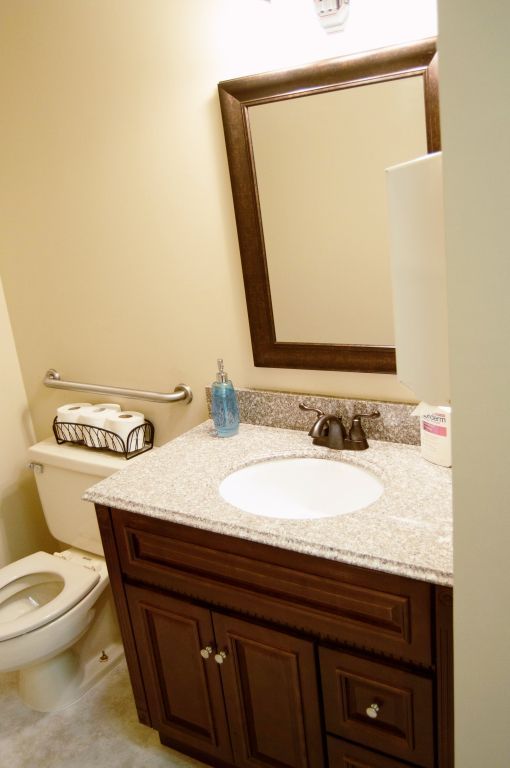 Bathroom with dark brown vanity, granite countertop, and toilet. Beige walls, a mirror, and a silver handrail.