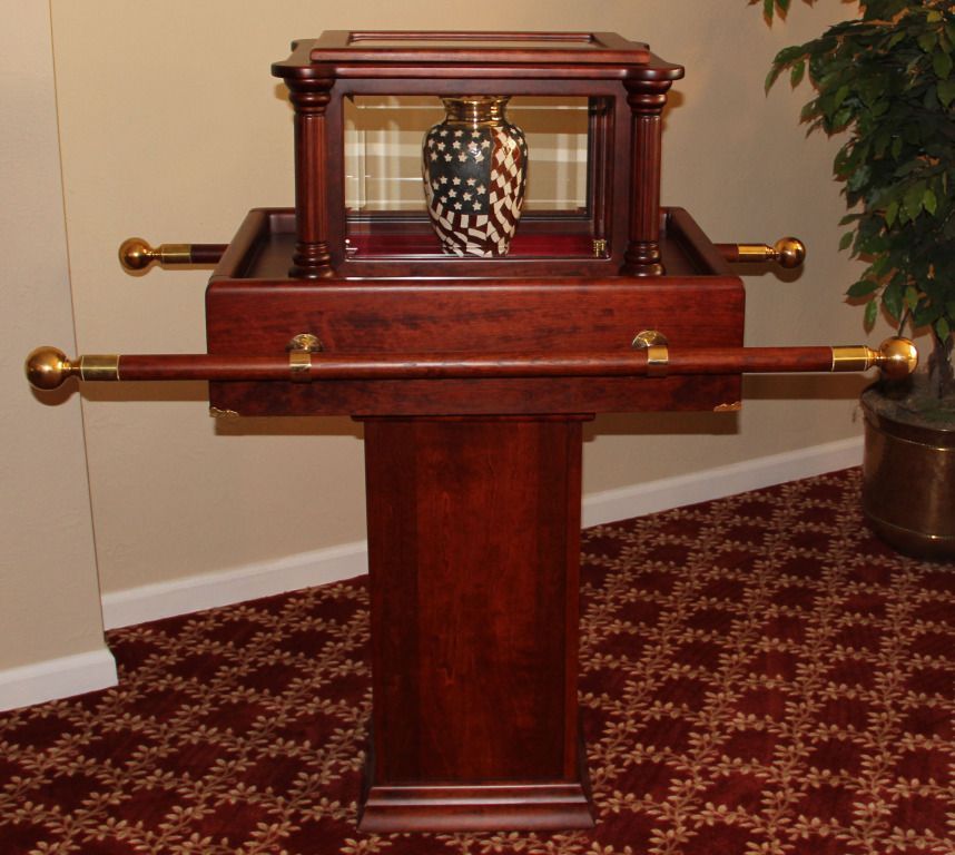 Wooden display stand with glass-enclosed urn. Brown, gold accents, and burgundy carpet.