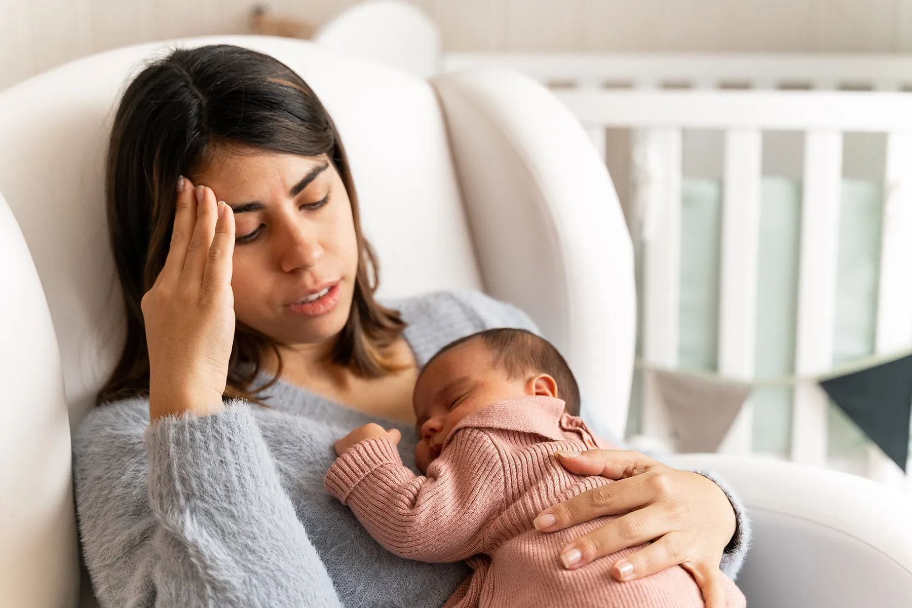 Tired new mother holding her baby while sitting in a nursery chair, reflecting postpartum fatigue 