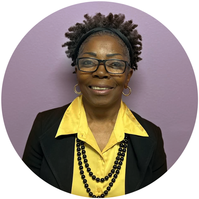 Woman with glasses, wearing a black blazer over a yellow shirt, with black beaded necklace, in front of purple background.