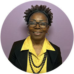 Woman with glasses, wearing a black blazer over a yellow shirt, with black beaded necklace, in front of purple background.