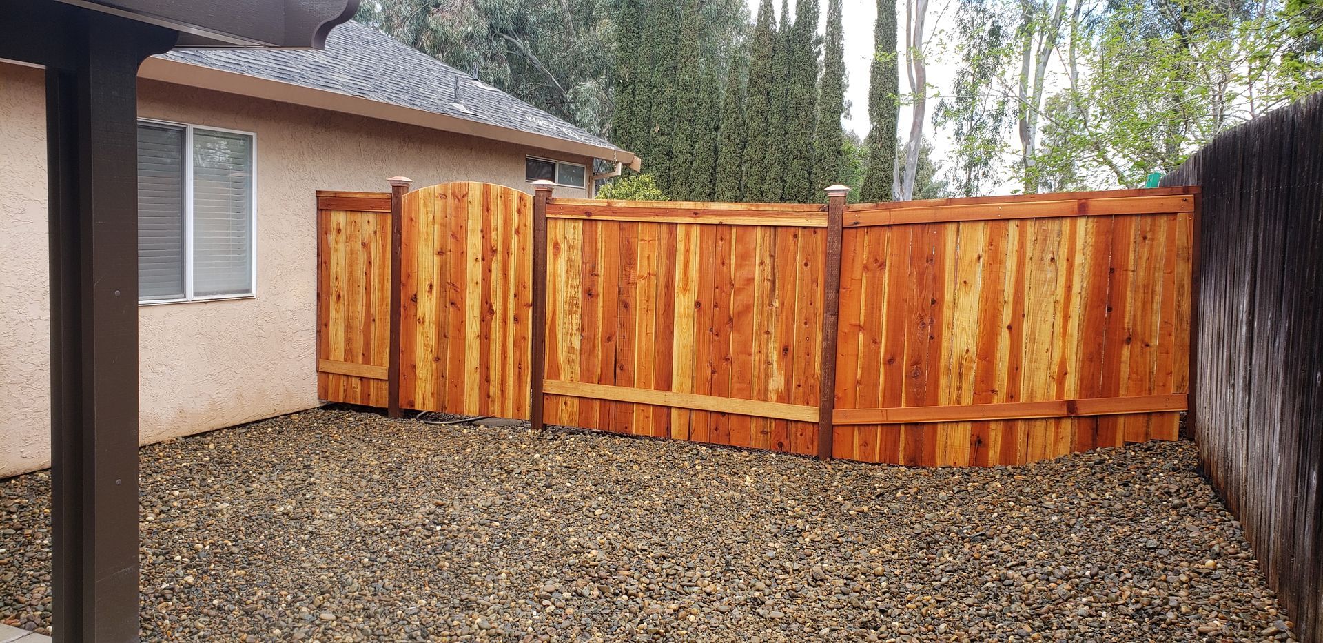 A new, stained wood fence with a gate stands in a backyard with a gravel ground surface next to a tan house.