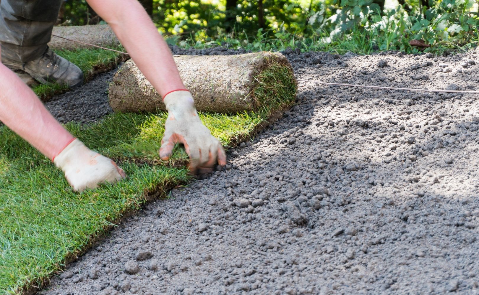 Person laying sod rolls on prepared soil outdoors.