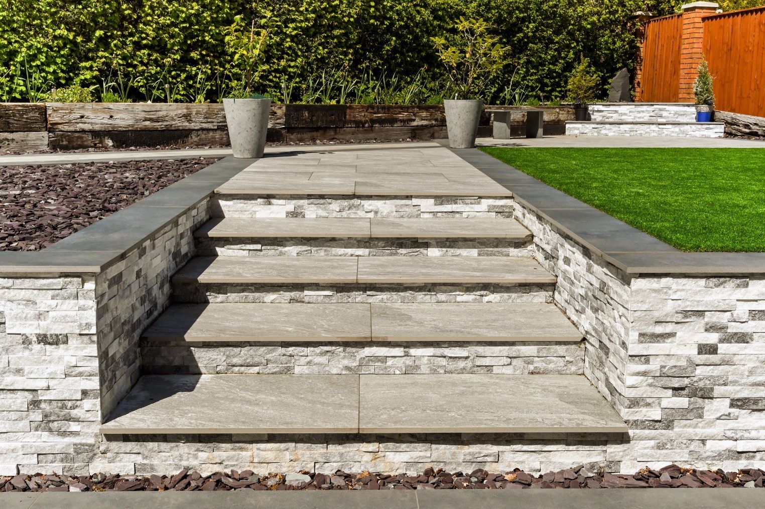 Stone steps leading up to a raised garden bed with two potted plants, surrounded by stone walls and green lawn.