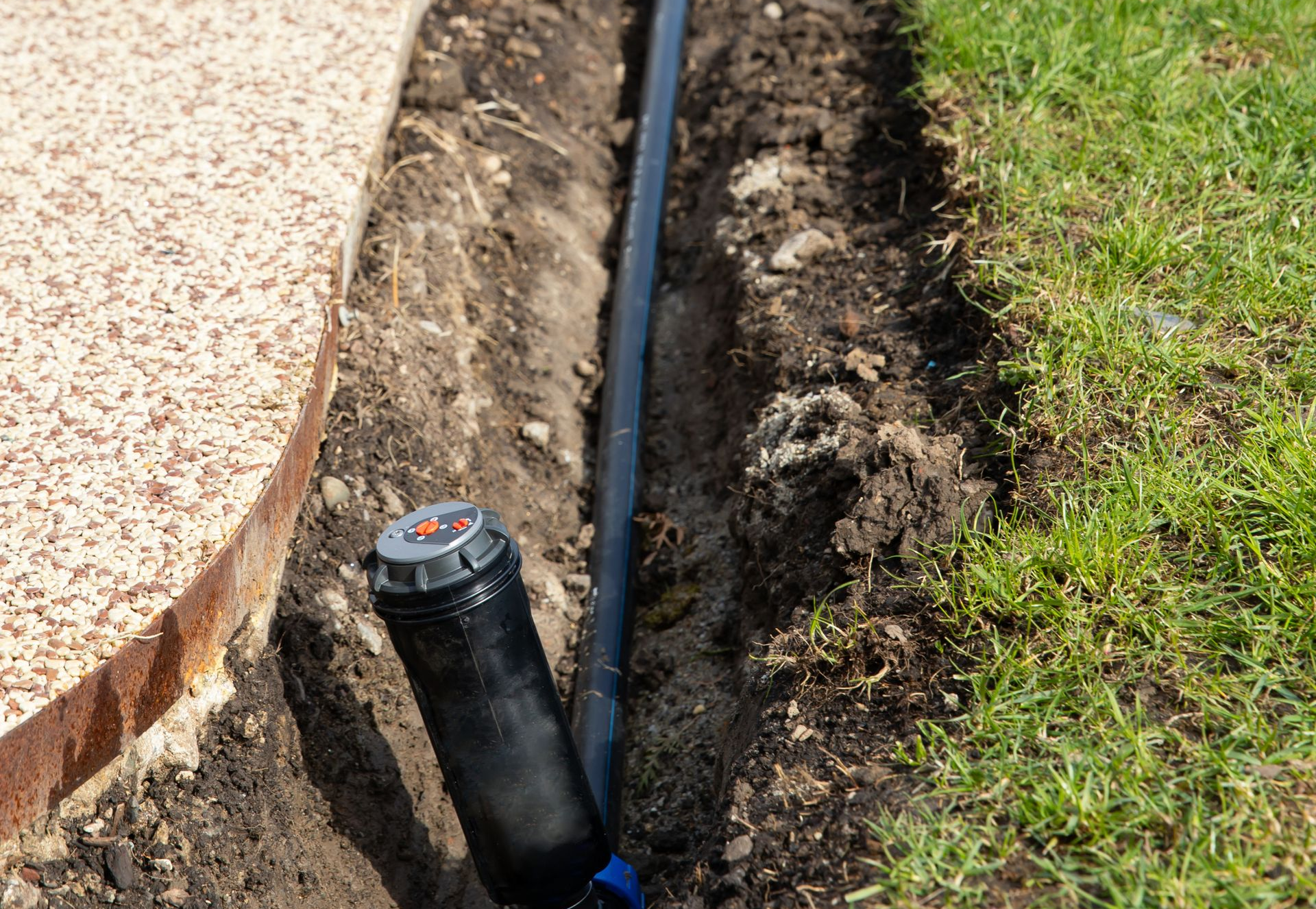 A black sprinkler head attached to a black irrigation pipe sits in a narrow trench next to a walkway and grass.