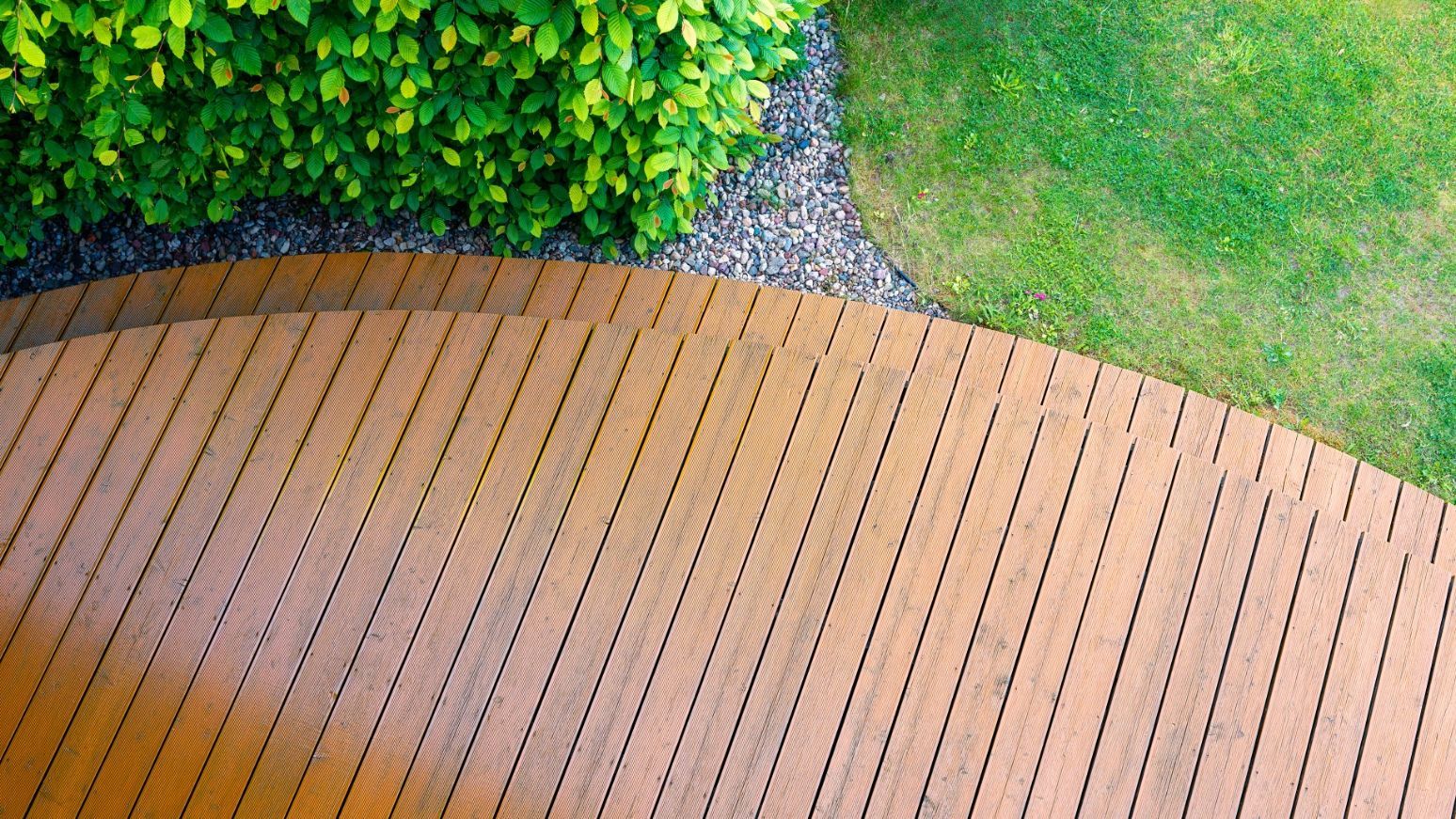 Overhead view: Wooden deck curving next to gravel, green grass, and leafy green hedge.