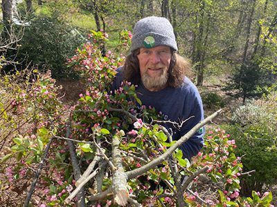 A man with long hair and a beard is standing next to a tree with pink flowers.