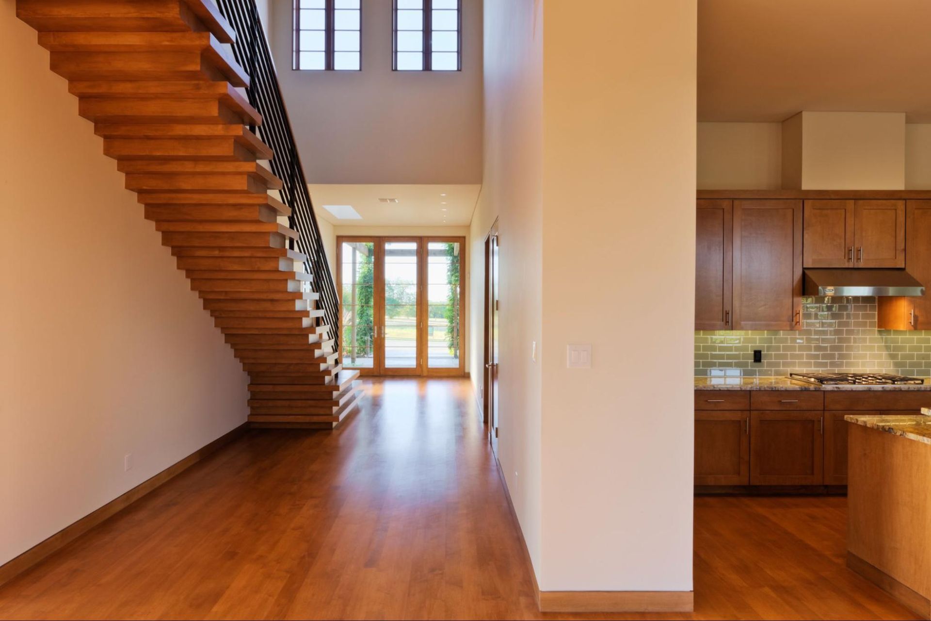 Modern home interior with a floating wooden staircase, polished hardwood floors, and a glimpse of a kitchen with wood cabinets.
