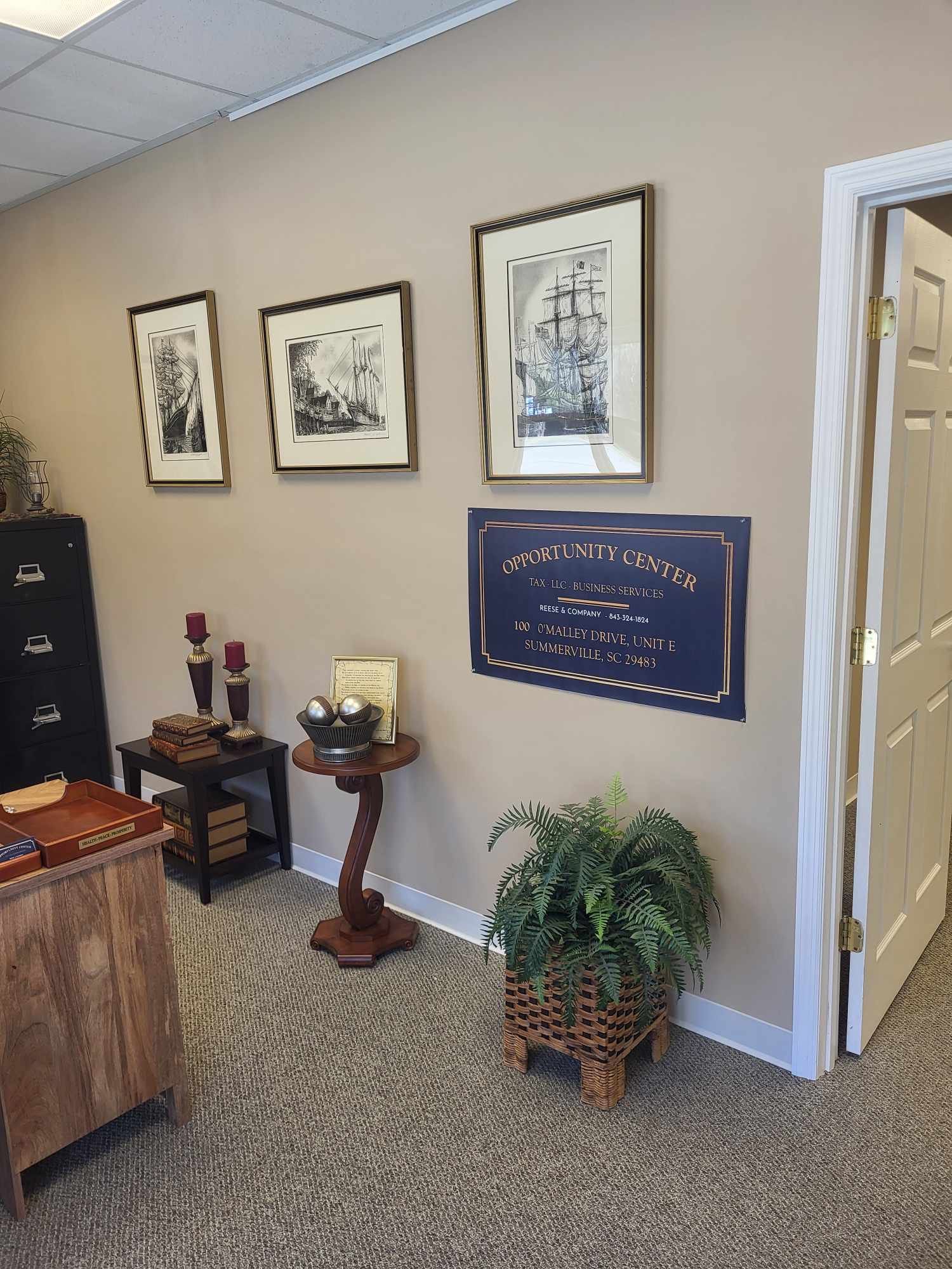 An office corner with framed art, potted plant, dark furniture, and a doorway. Beige walls and carpet.