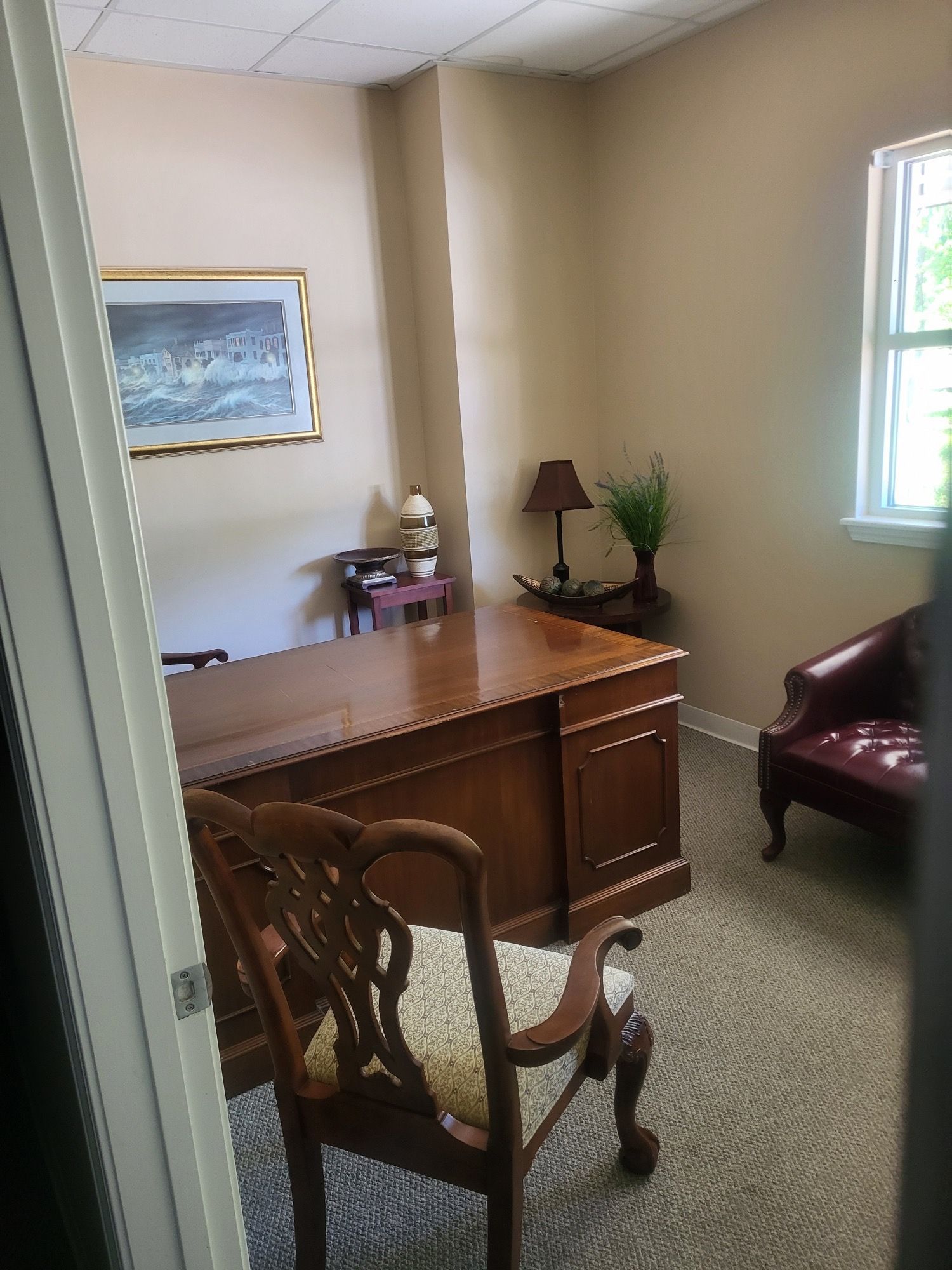 Office with a large wooden desk and chair, red chair, and artwork. Beige walls, window to the right.
