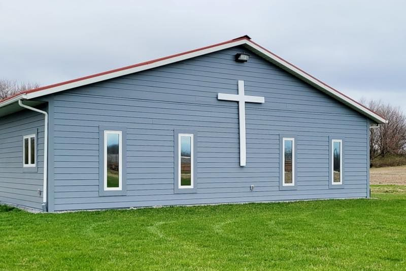 Red brick church with white trim and entrance. A sign stands in front on the green lawn.