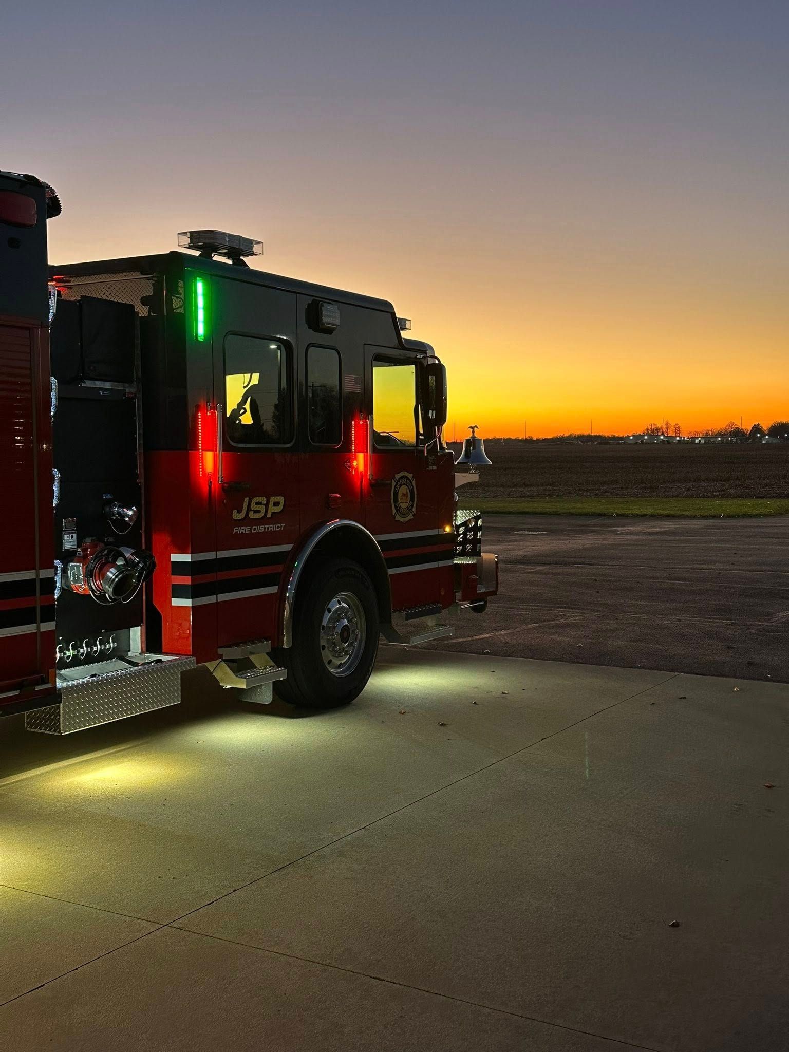 Fire truck parked on pavement at sunset. Red and black truck, green and red lights on.
