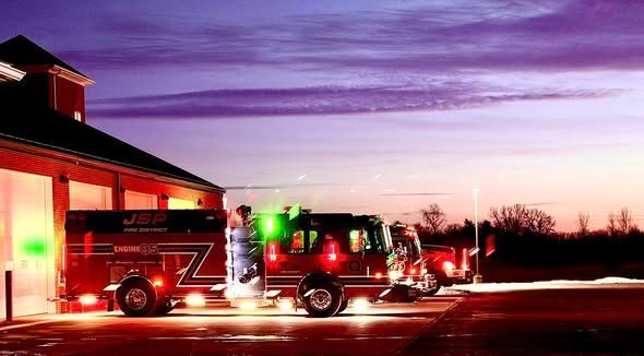 Fire trucks parked outside a station at dusk, illuminated with green and red lights.