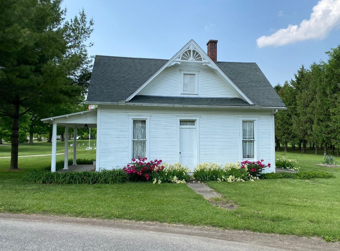 White house with porch, gabled roof, and decorative trim, set on green lawn with flowering plants.