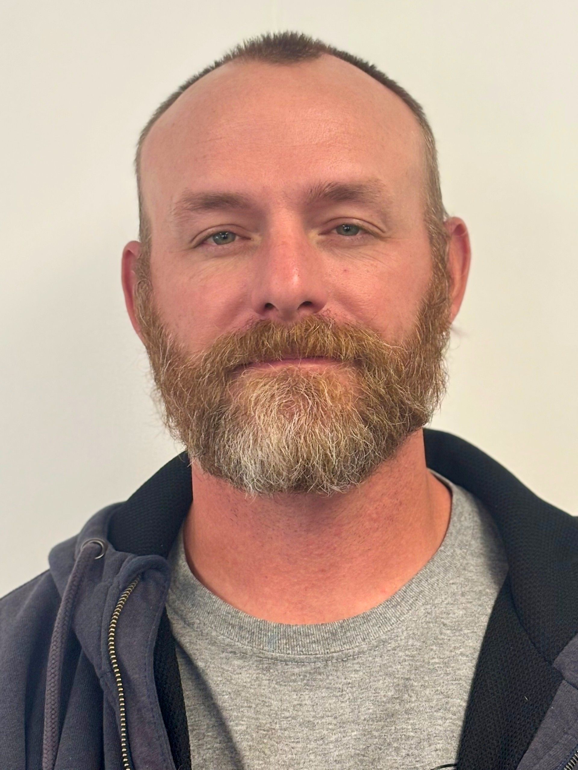 Man with a brown beard and short hair wearing a gray shirt and dark blue zip-up jacket.
