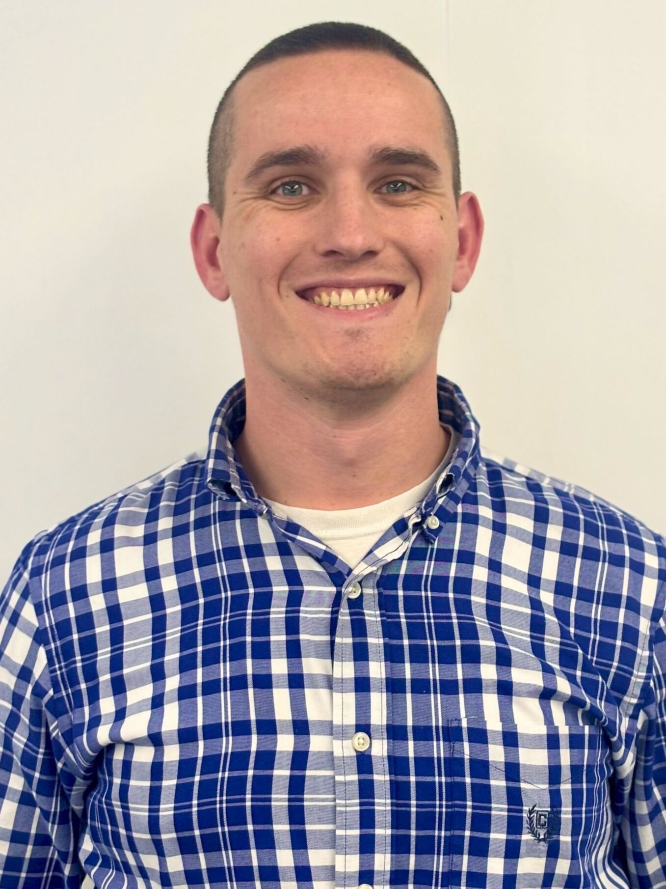 Man with a shaved head and blue plaid shirt smiling, against a white backdrop.