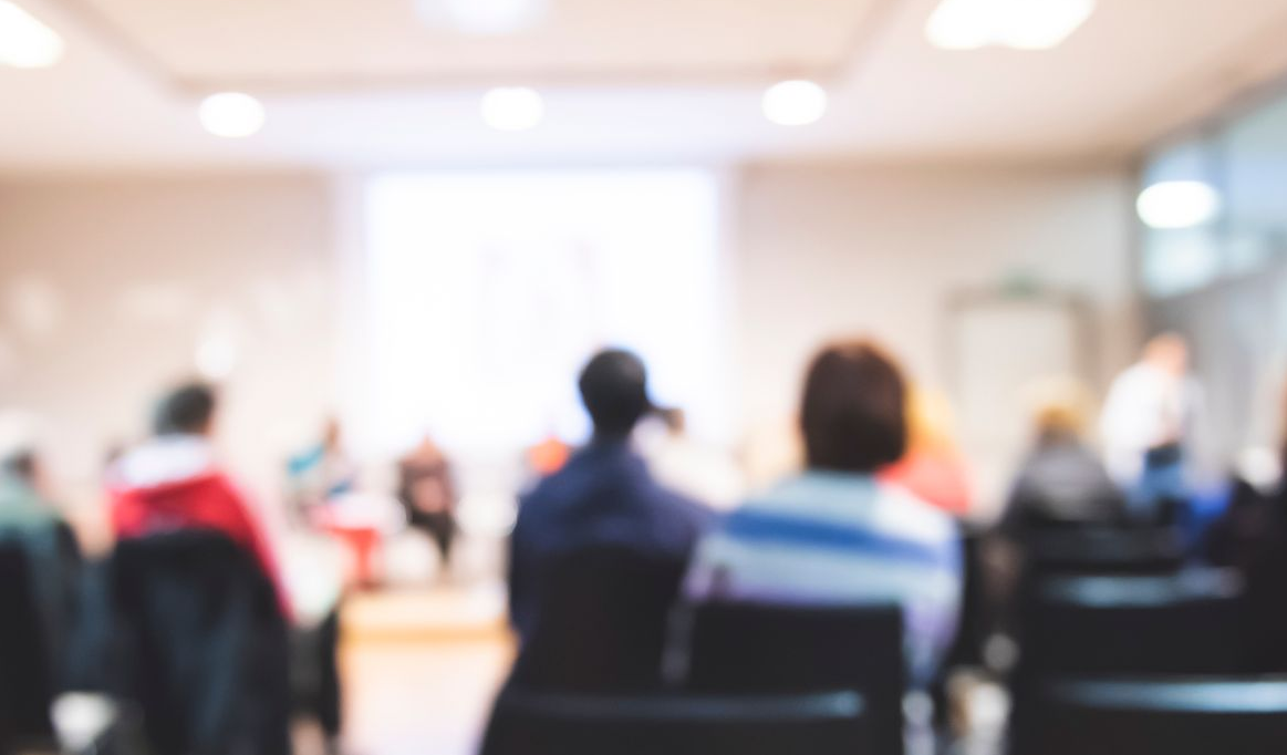 Blurred view of an audience in a presentation room, focused on backs of heads, bright screen in background.