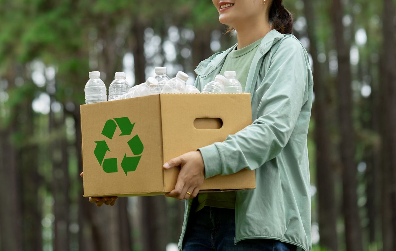 Person smiling, carrying a cardboard box with a recycling symbol, filled with plastic bottles, in a park.