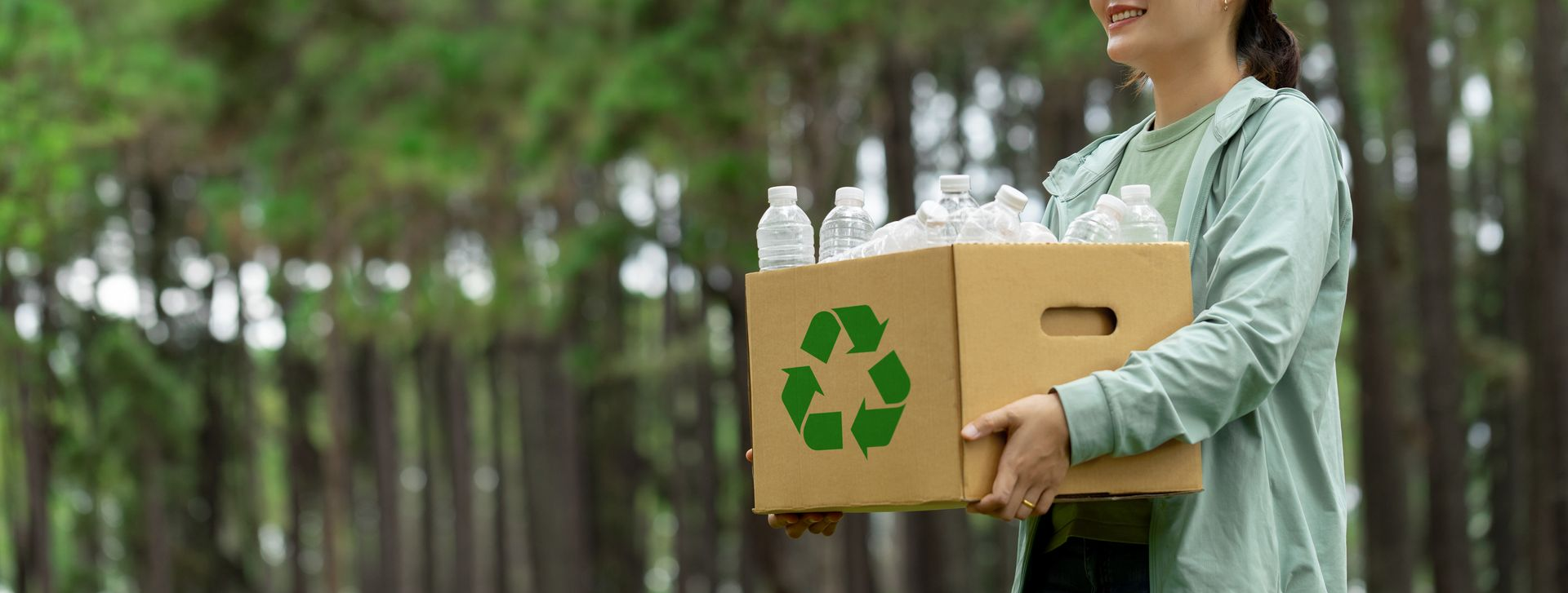 Person carrying a cardboard box with recycling symbol and plastic bottles in a green forest setting.
