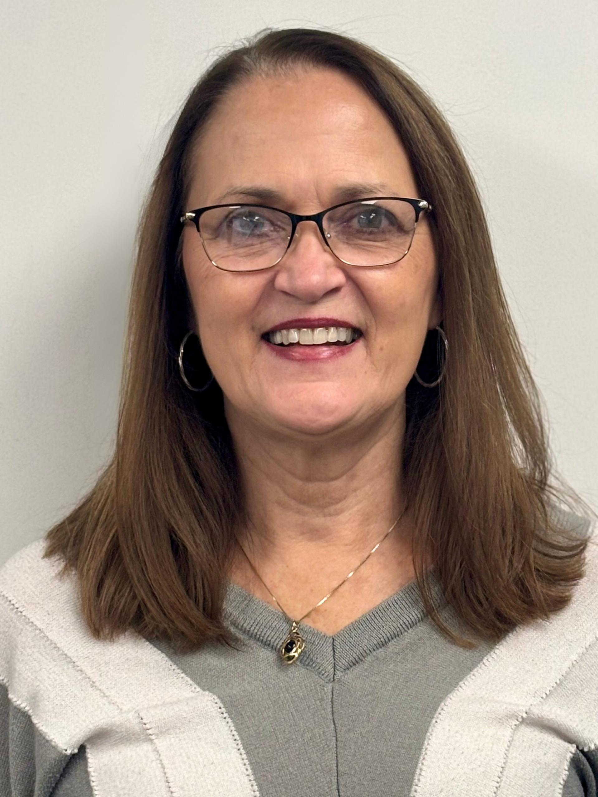 Woman with glasses and a smiling expression, wearing a gray and white top, standing against a white wall.