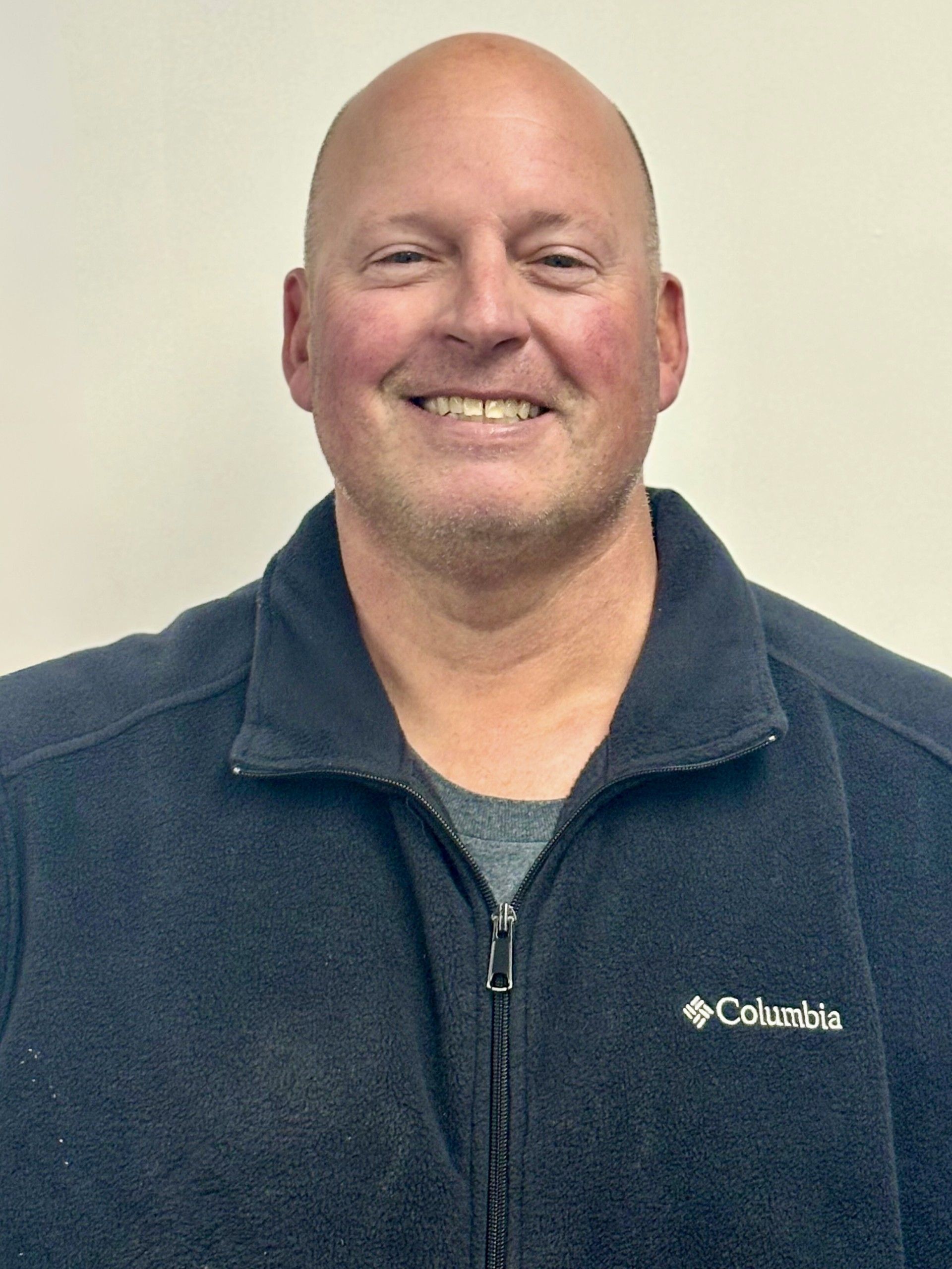 Bald man smiling, wearing a dark blue Columbia fleece jacket in front of a neutral wall.