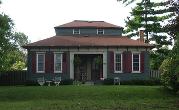 Green house with brown roof, shutters, and small garden with chairs on a green lawn.