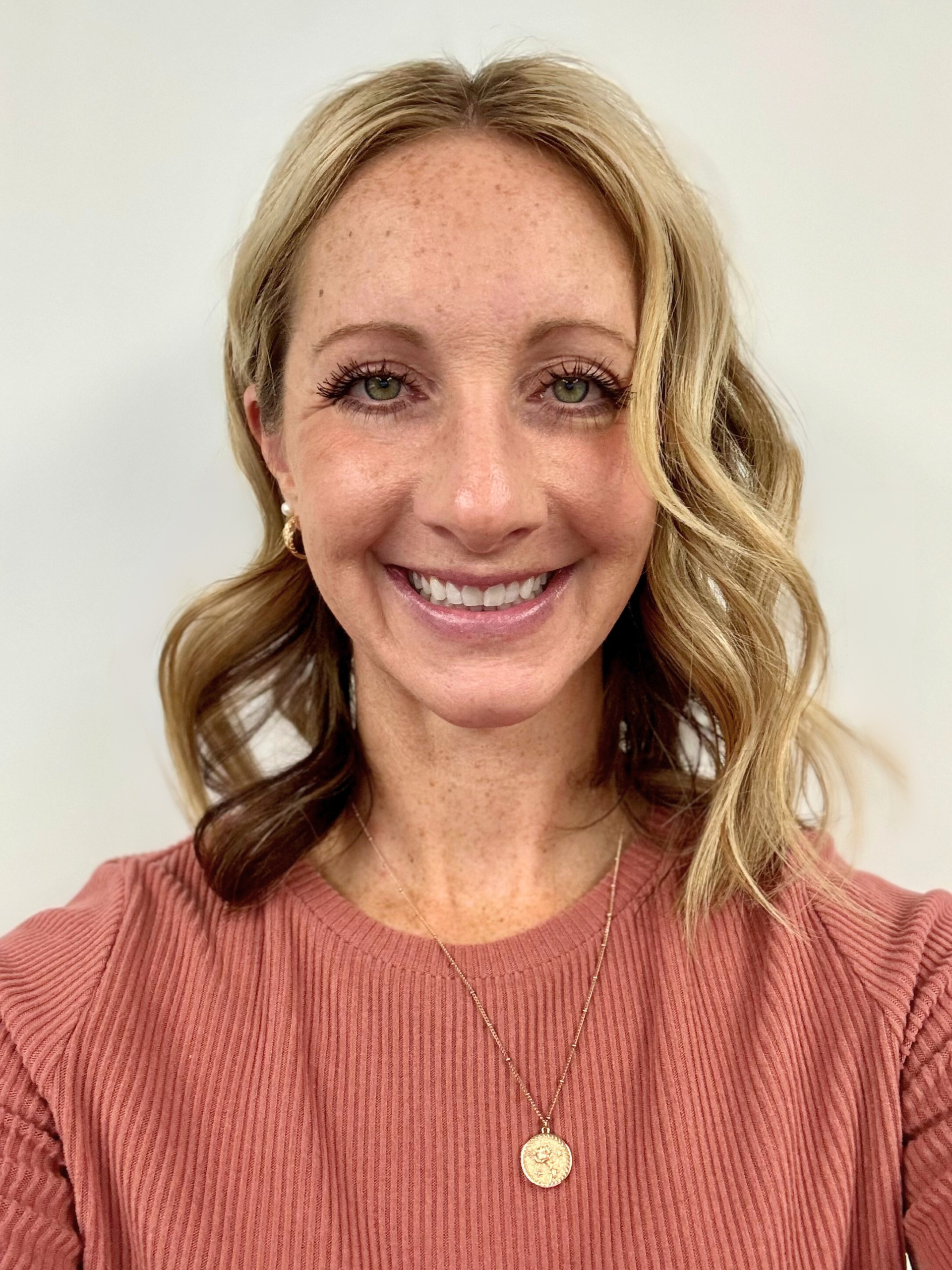 Woman with wavy blonde hair, freckles, and a wide smile, wearing a pink shirt and necklace, posing in front of a white wall.