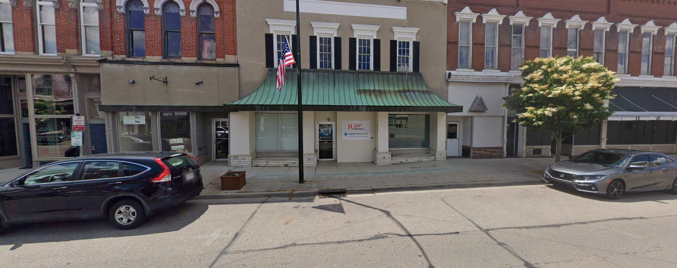 Buildings on a street, a black SUV parked on the left, and a gray car on the right. An American flag hangs on the building in the middle.