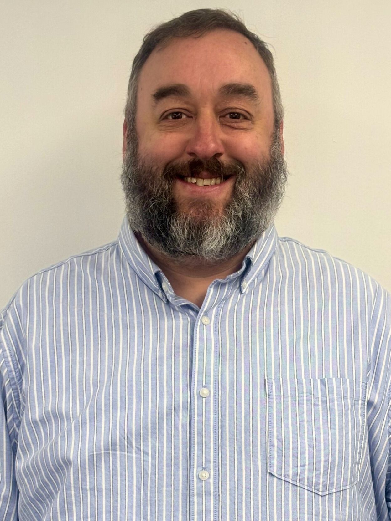 Man with beard smiles, wearing a blue and white striped shirt, against a white wall.