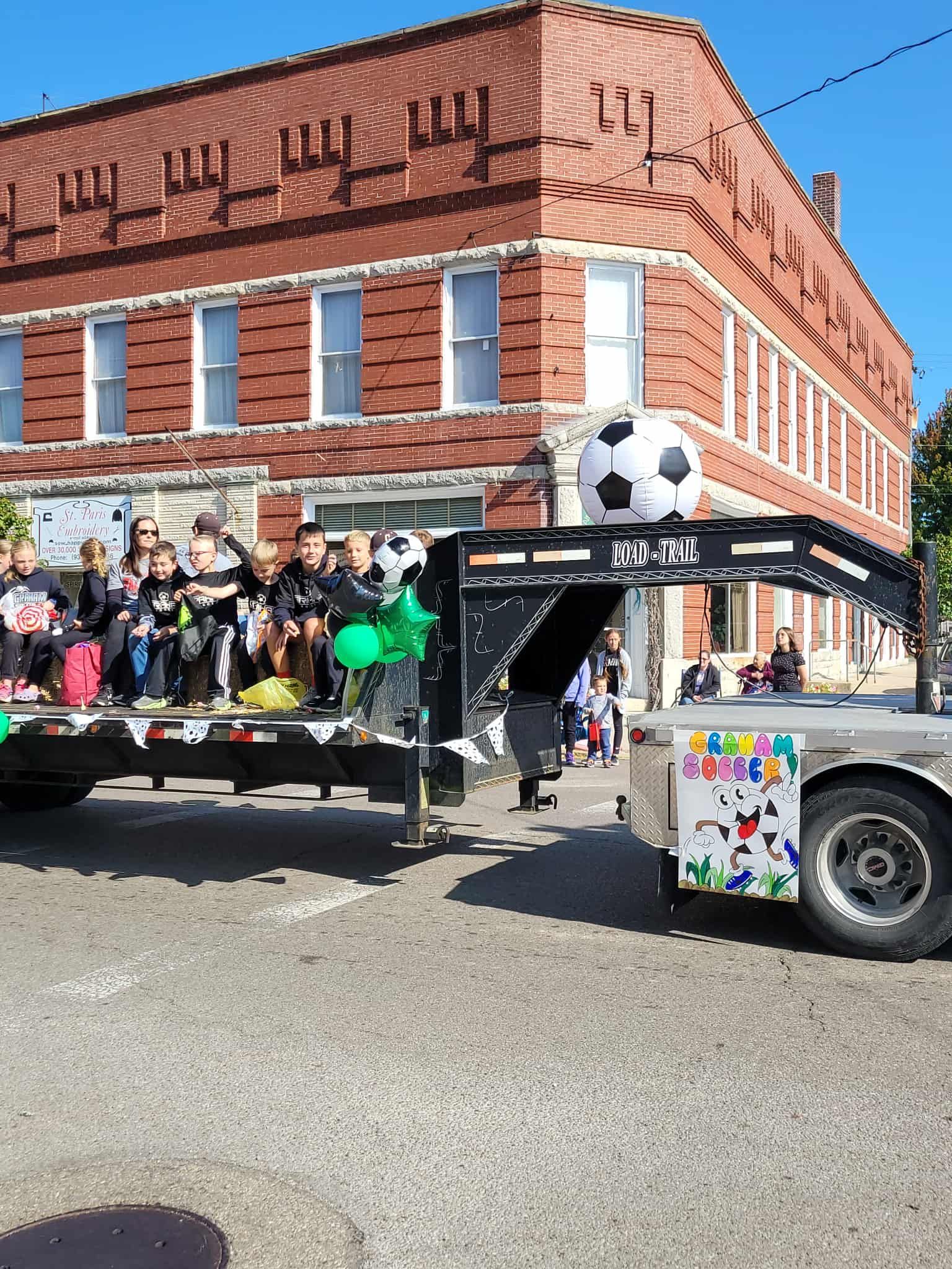 Soccer team on a flatbed trailer in a parade, soccer ball on top, spectators in front of brick building.