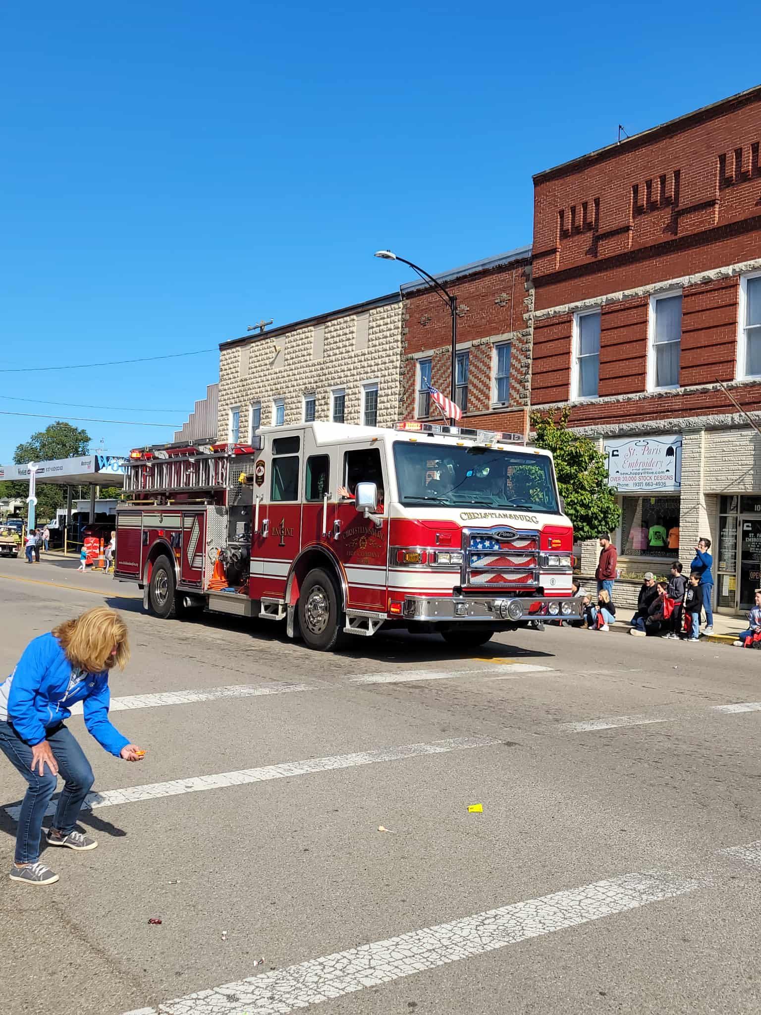Fire truck in a parade, driving past brick buildings, a person on the left picks something up off the street.
