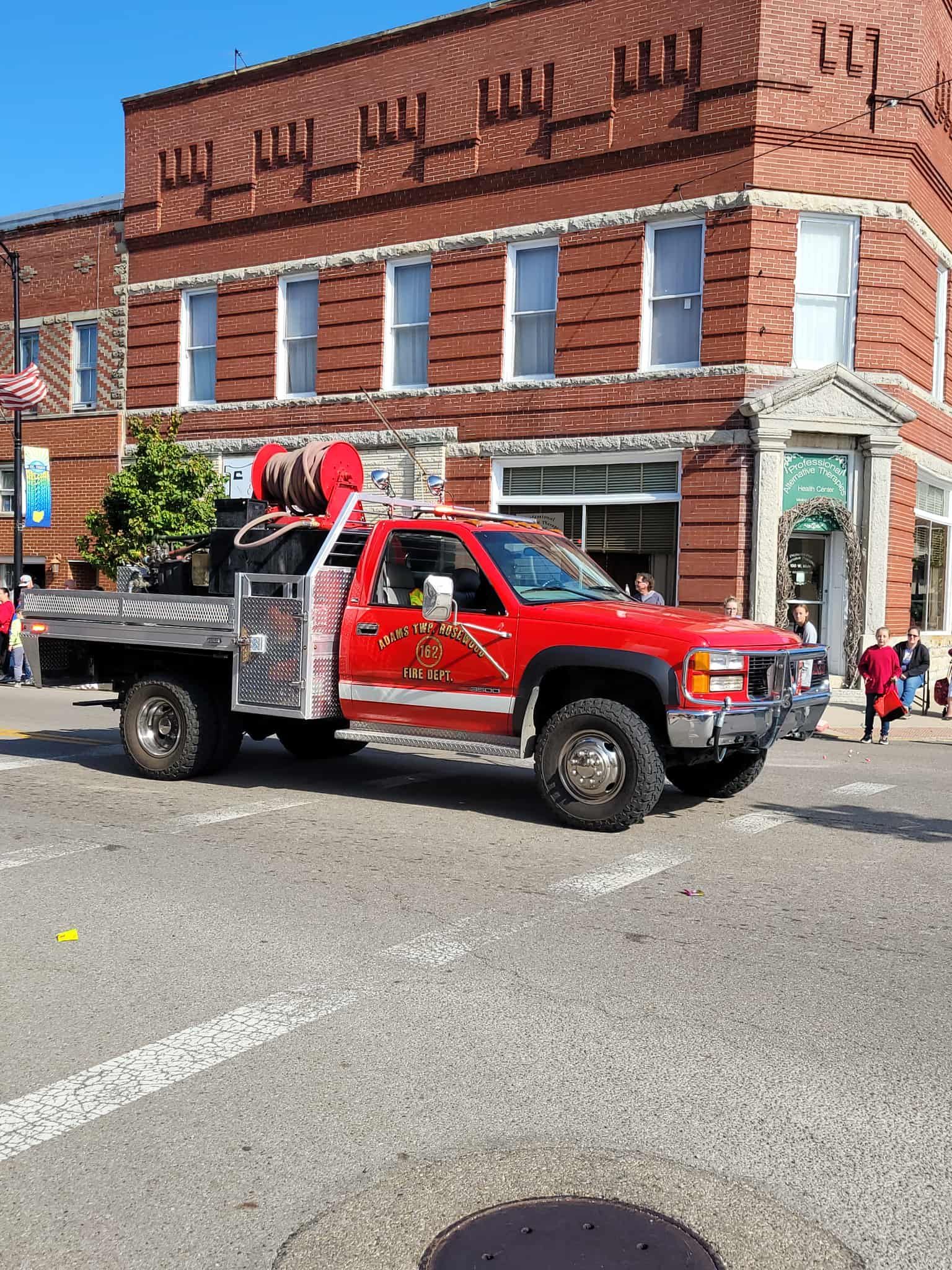 Red fire department truck in a parade, driving past a red brick building with white trim.