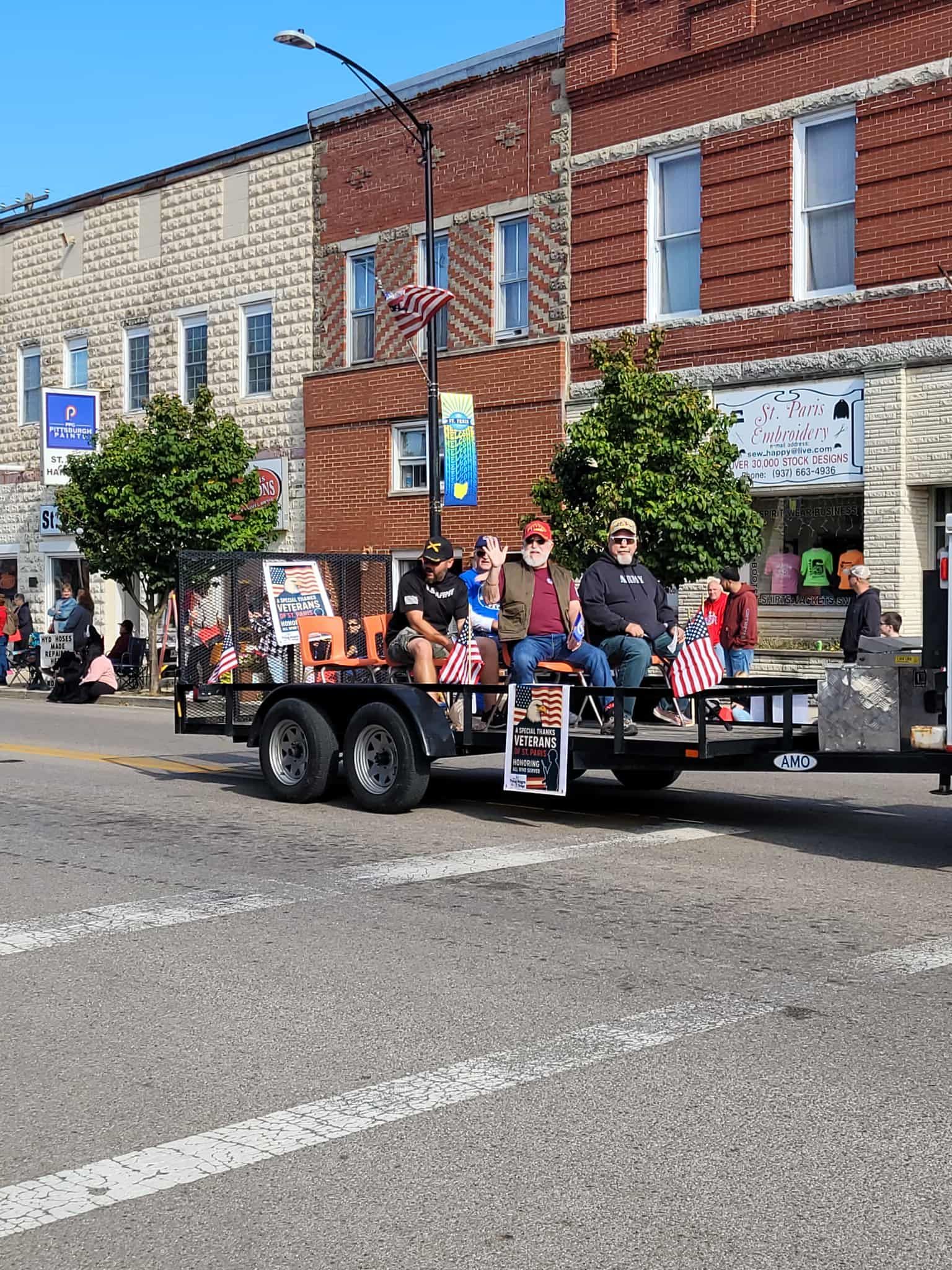People on a trailer float in a parade, passing buildings.